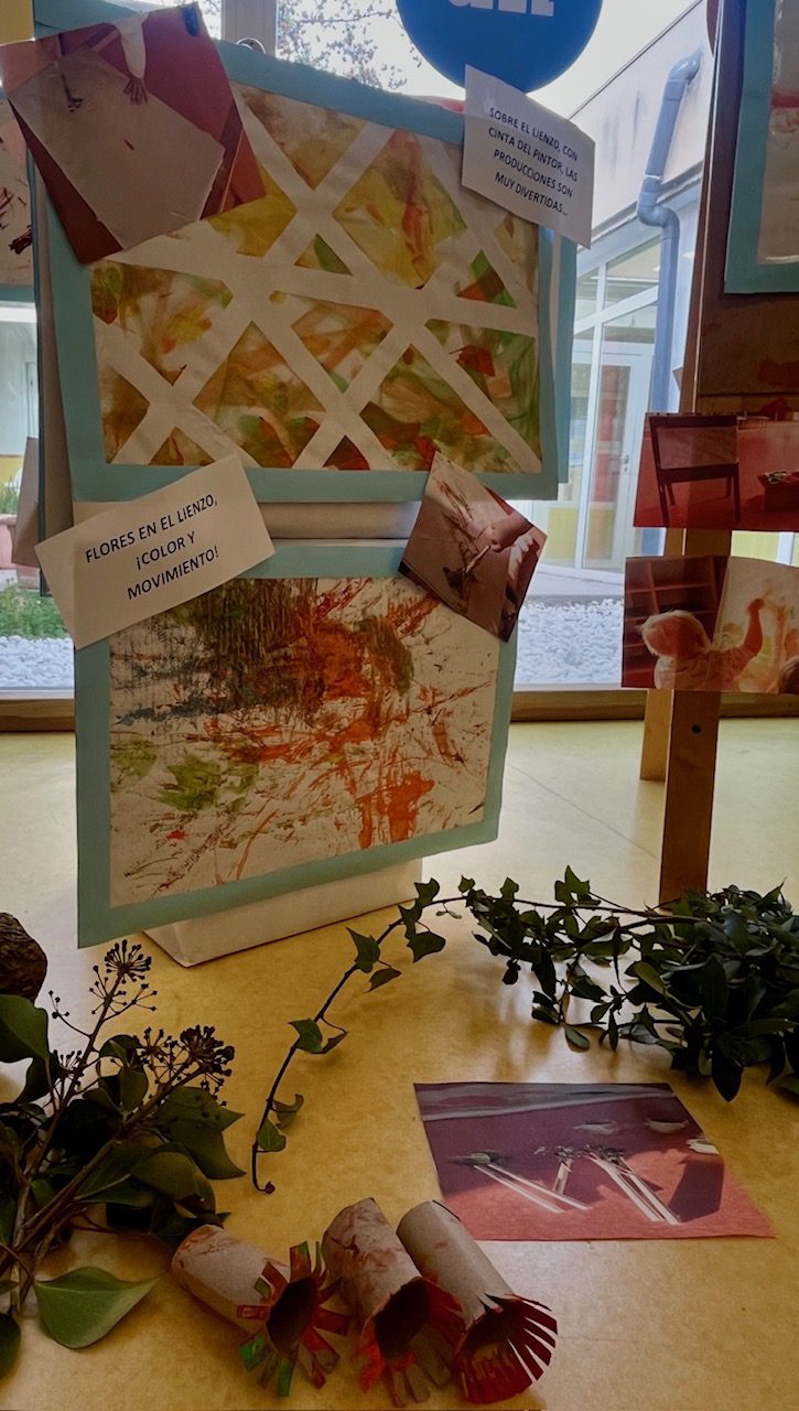 Detalle de pinturas infantiles hechas con cintas, flores y sellos de cartón reciclado en el suelo de la escuela.